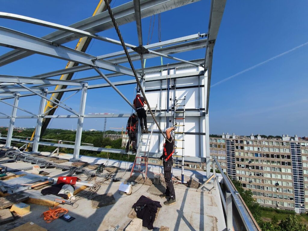 Bouwvakkers werken aan een metalen constructie op hoogte, met een stedelijke omgeving op de achtergrond.