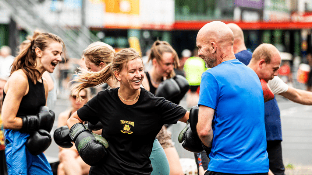Groep mensen doet enthousiast mee aan een bokstraining in de buitenlucht, lachend en actief bezig met elkaar.
