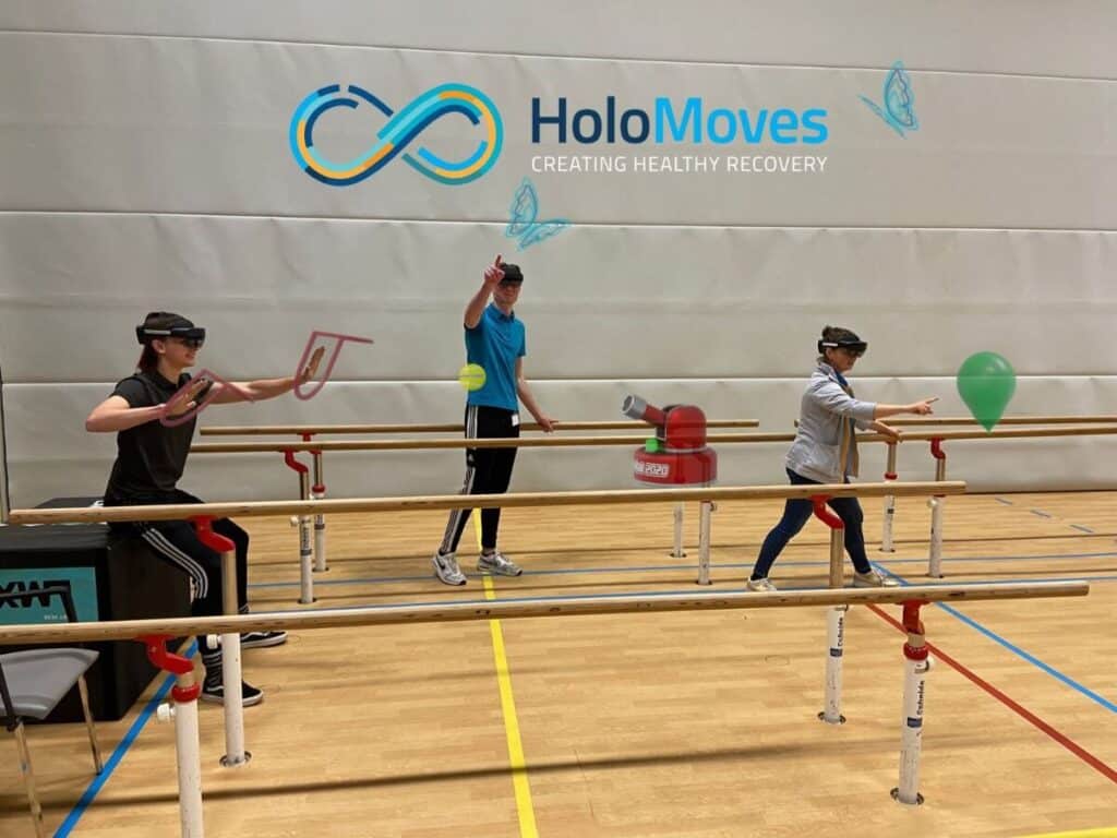 Drie mensen dragen VR-headsets en voeren oefeningen uit in een sporthal, omringd door virtuele objecten, onder een logo van HoloMoves.