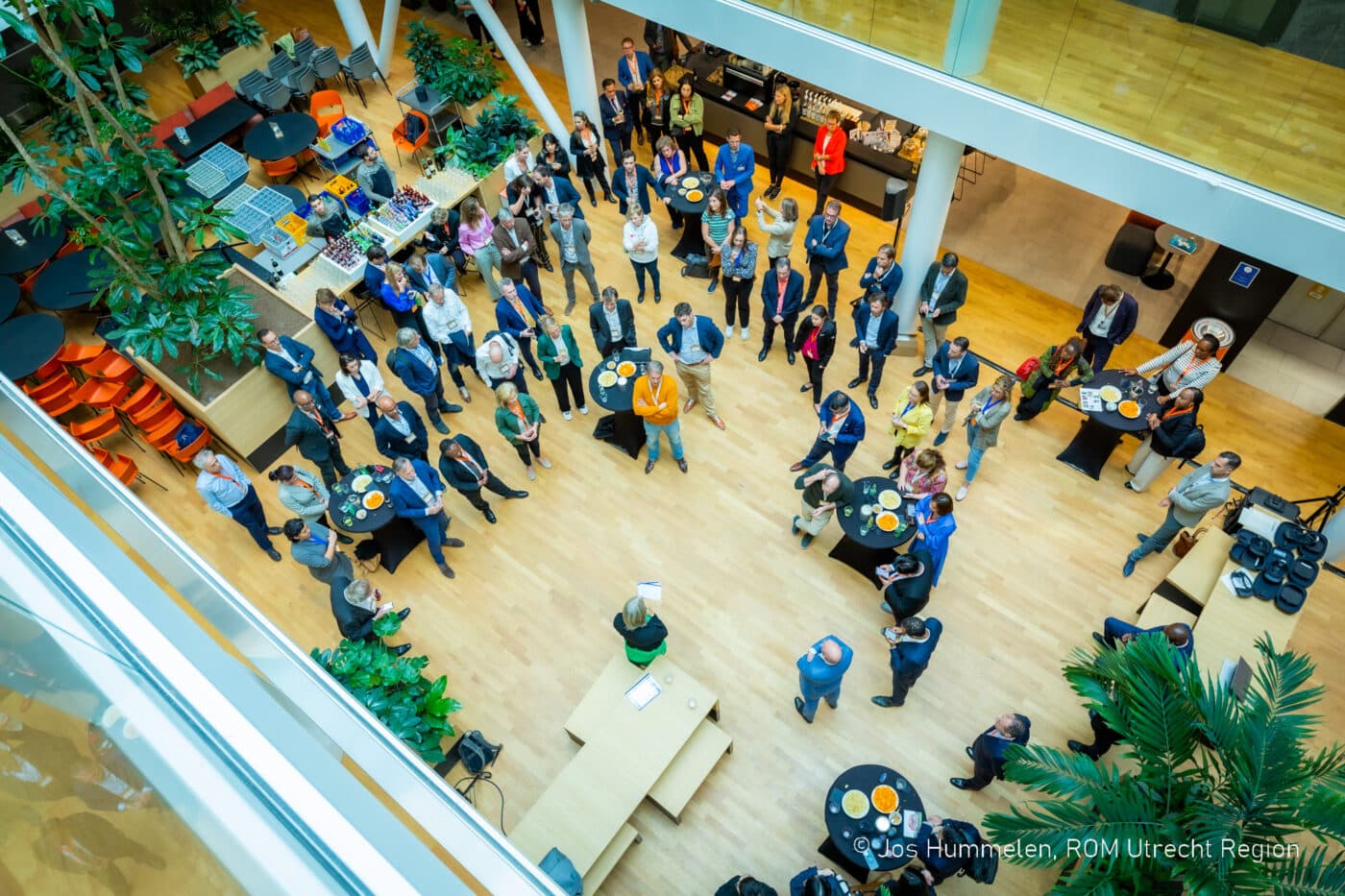 Presentatie van een evenement in een atrium, gezien van bovenaf, met ronde tafels en planten verspreid door de ruimte.