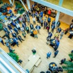 Presentatie van een evenement in een atrium, gezien van bovenaf, met ronde tafels en planten verspreid door de ruimte.